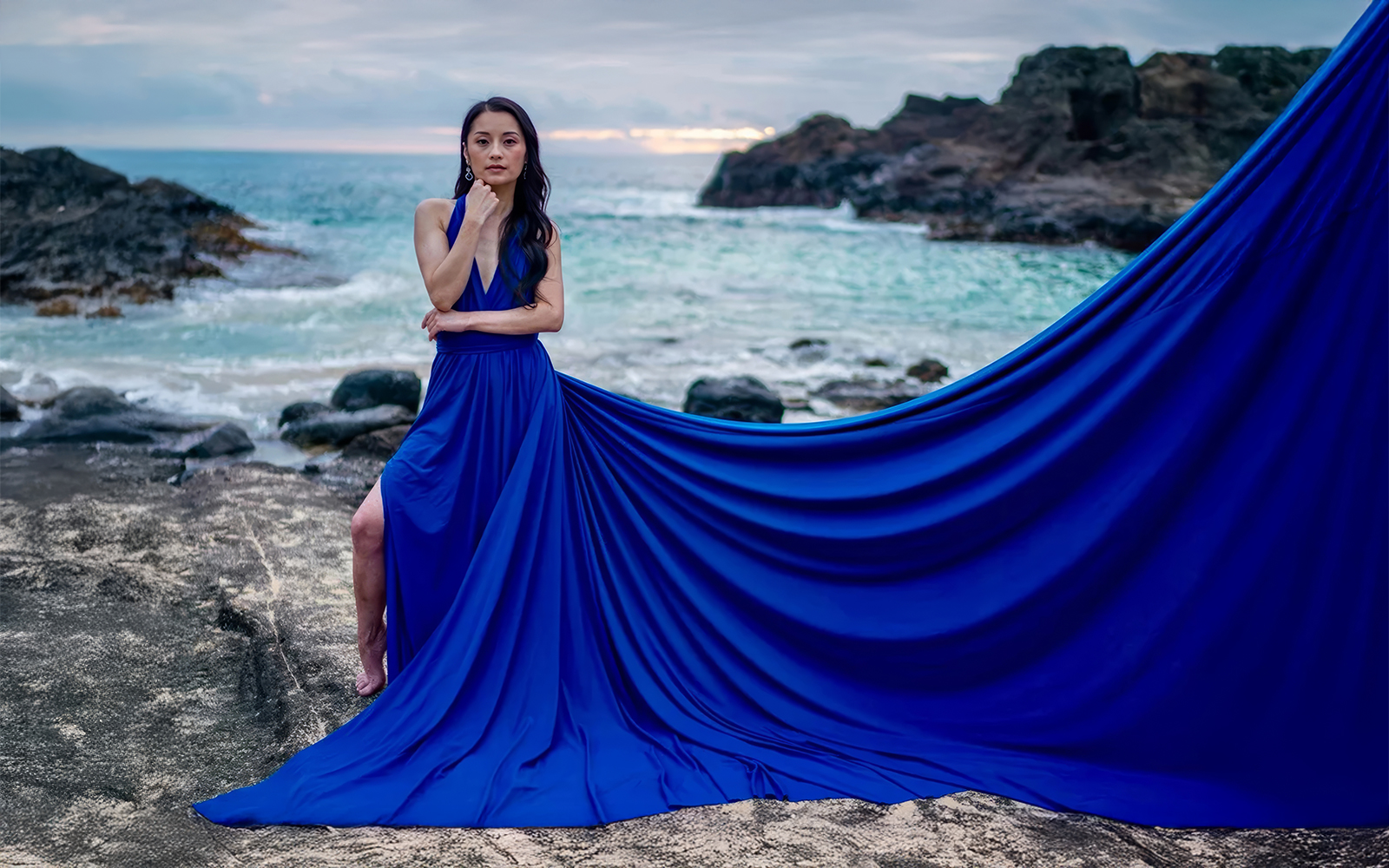 Woman in a royal blue flying dress with a long train across coastal rocks