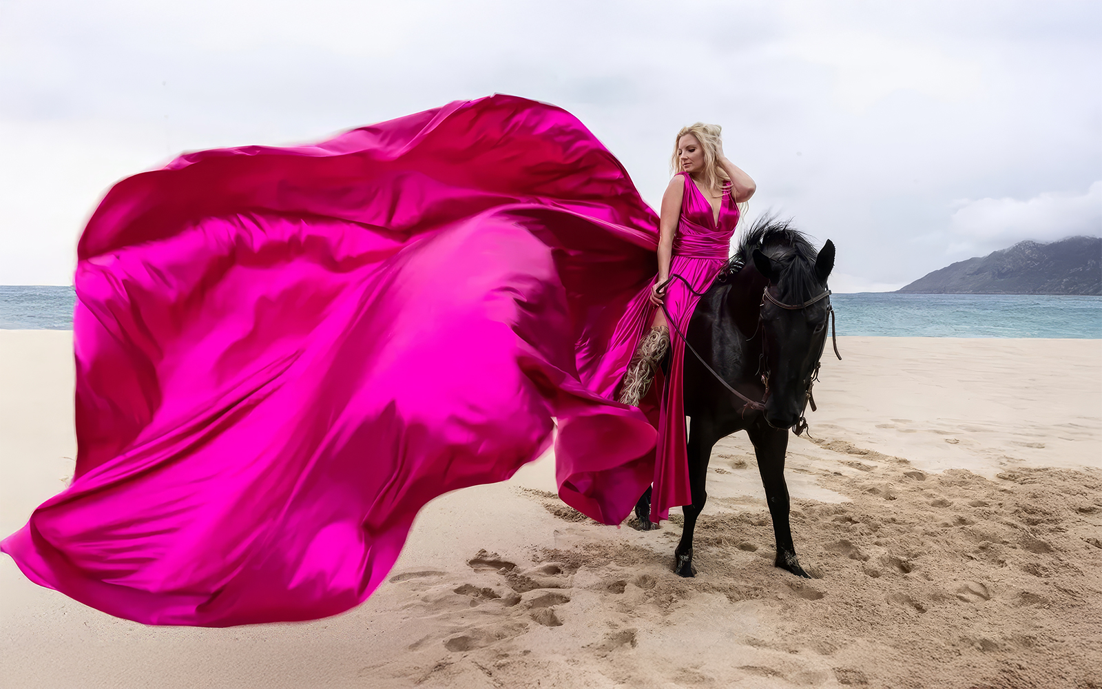 Woman in a glam pink flying dress on a beach beside a black horse