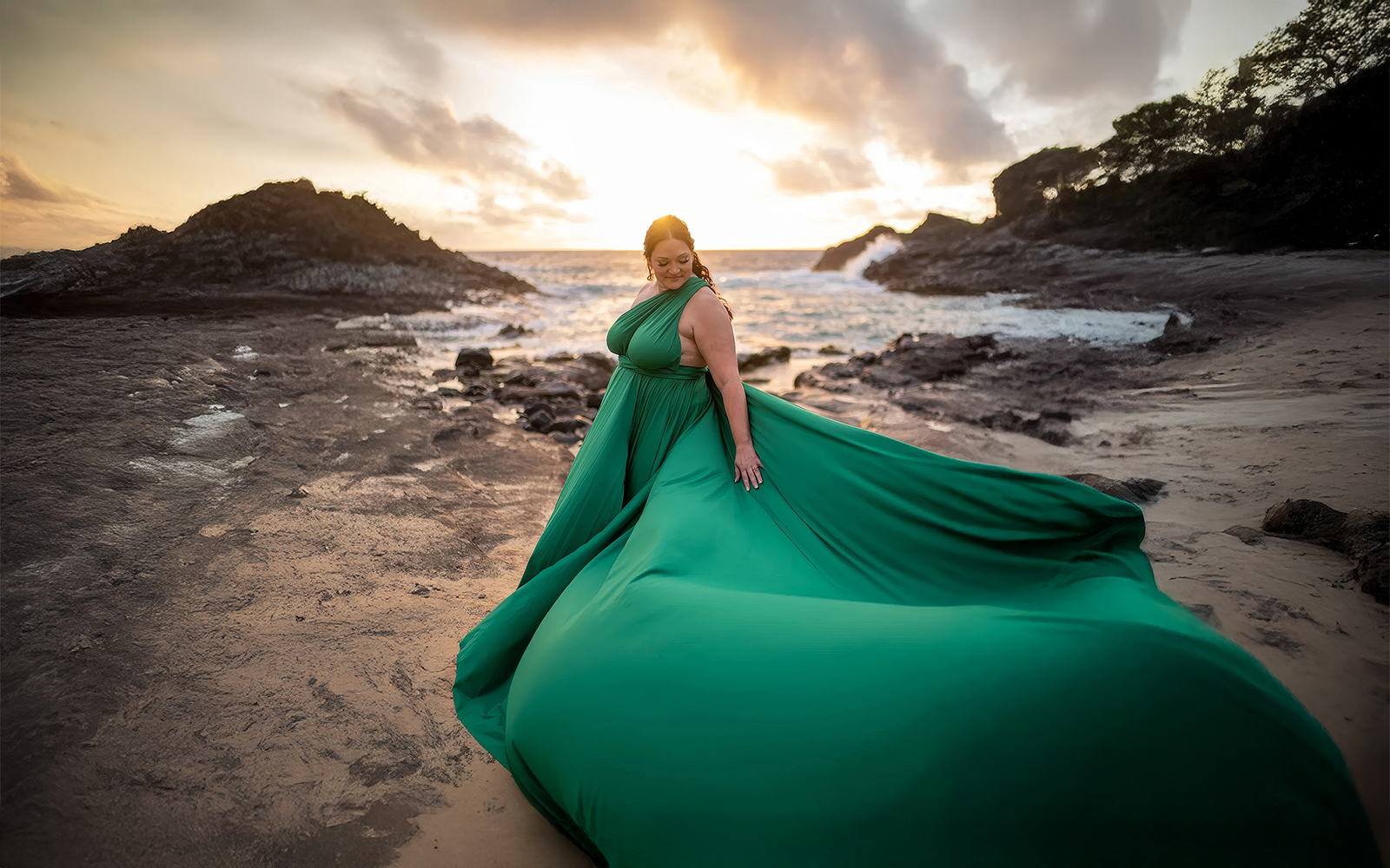 Woman in an emerald green flying dress on a rocky beach at sunset
