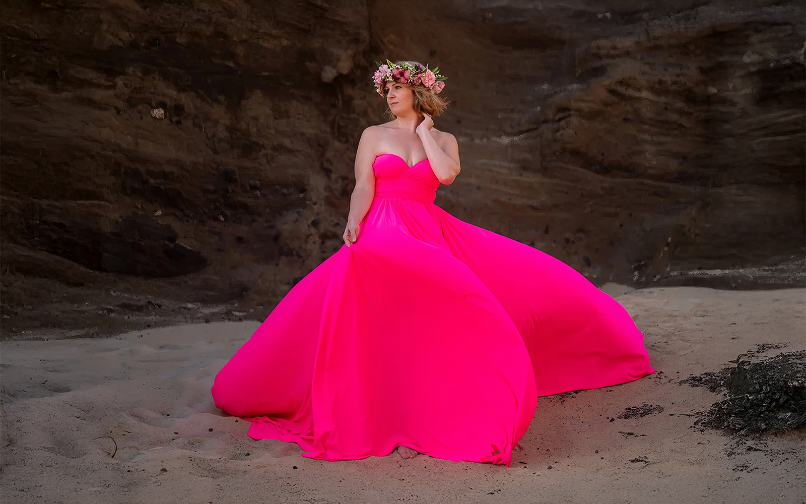 Woman in a hot pink flying dress on a beach in front of a rocky cliff