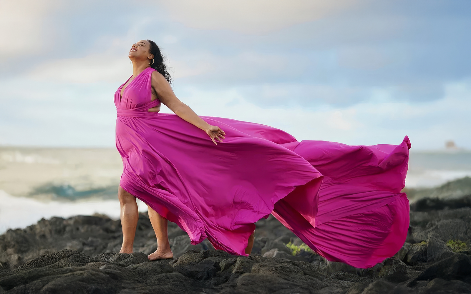 Woman in a magenta flying dress standing on coastal rocks by the sea