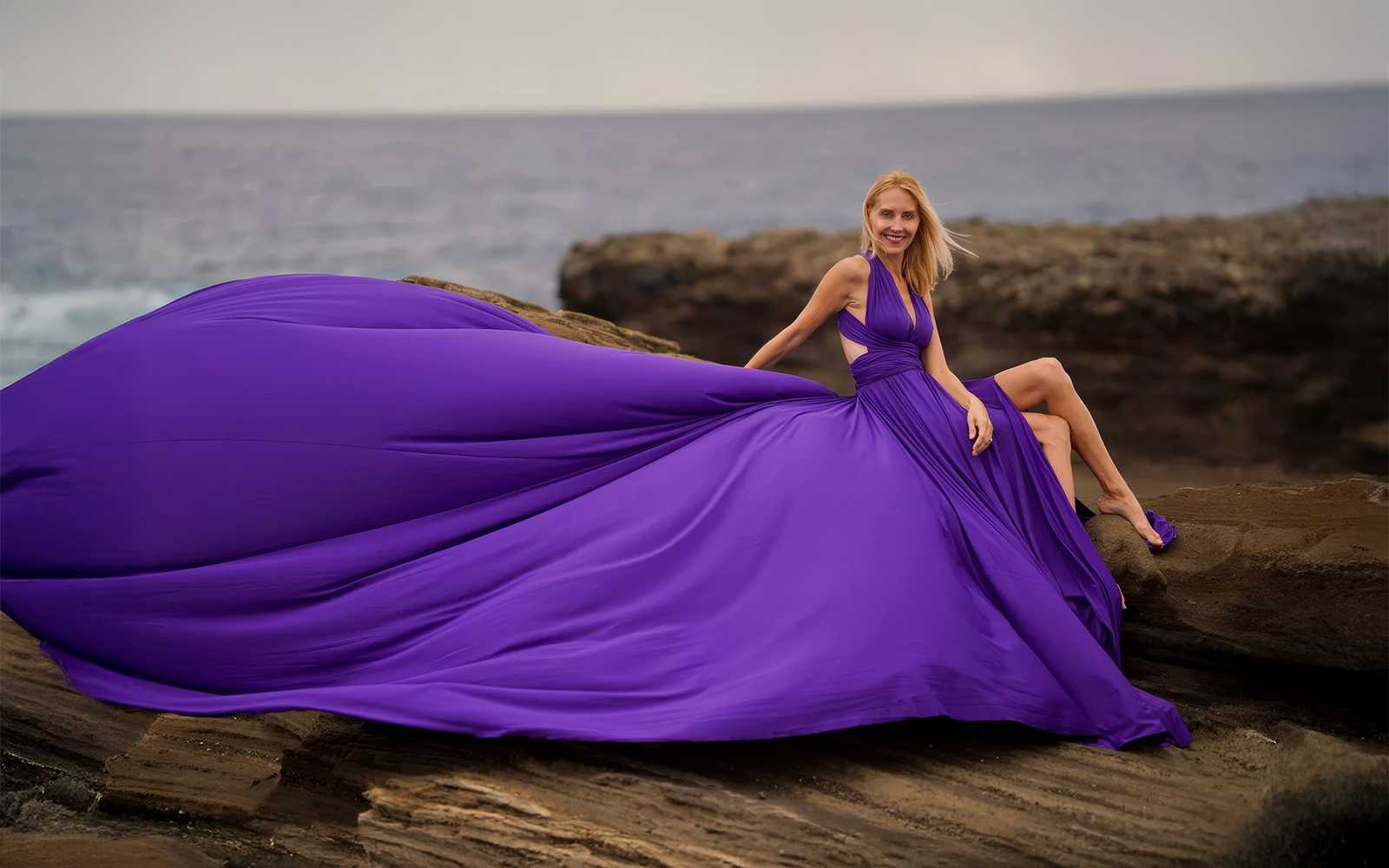 Woman reclining on shoreline rocks in a purple flying dress