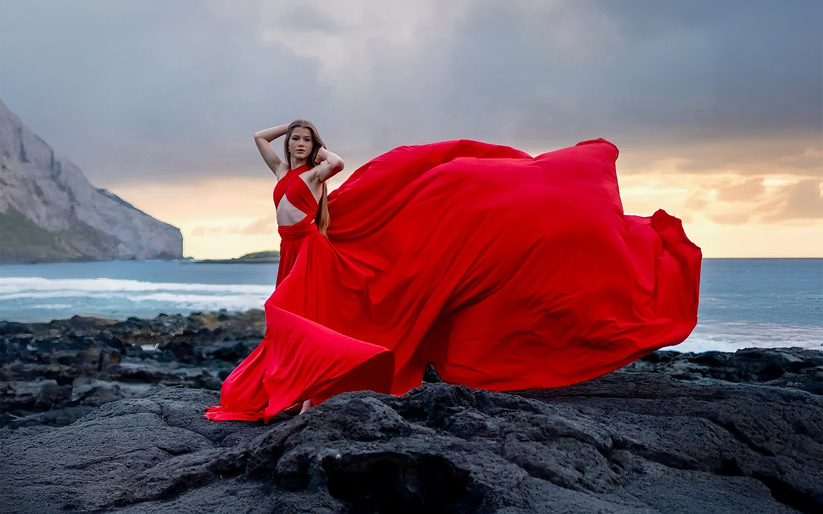 Woman in a red flying dress on dark lava rocks at the ocean's edge
