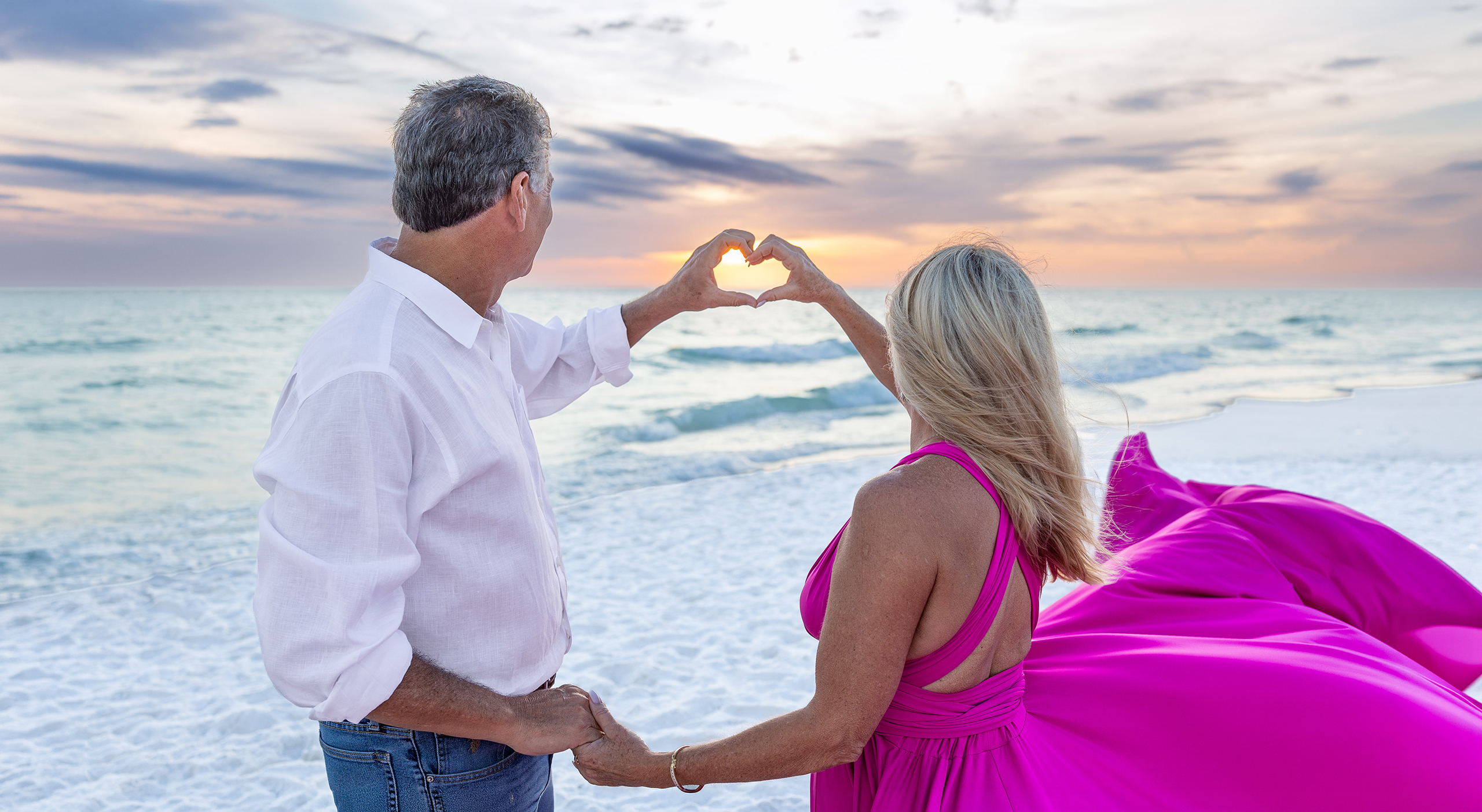 SUNSET~2 Couple making a heart shape with their hands at sunset, the woman in a magenta flying dress