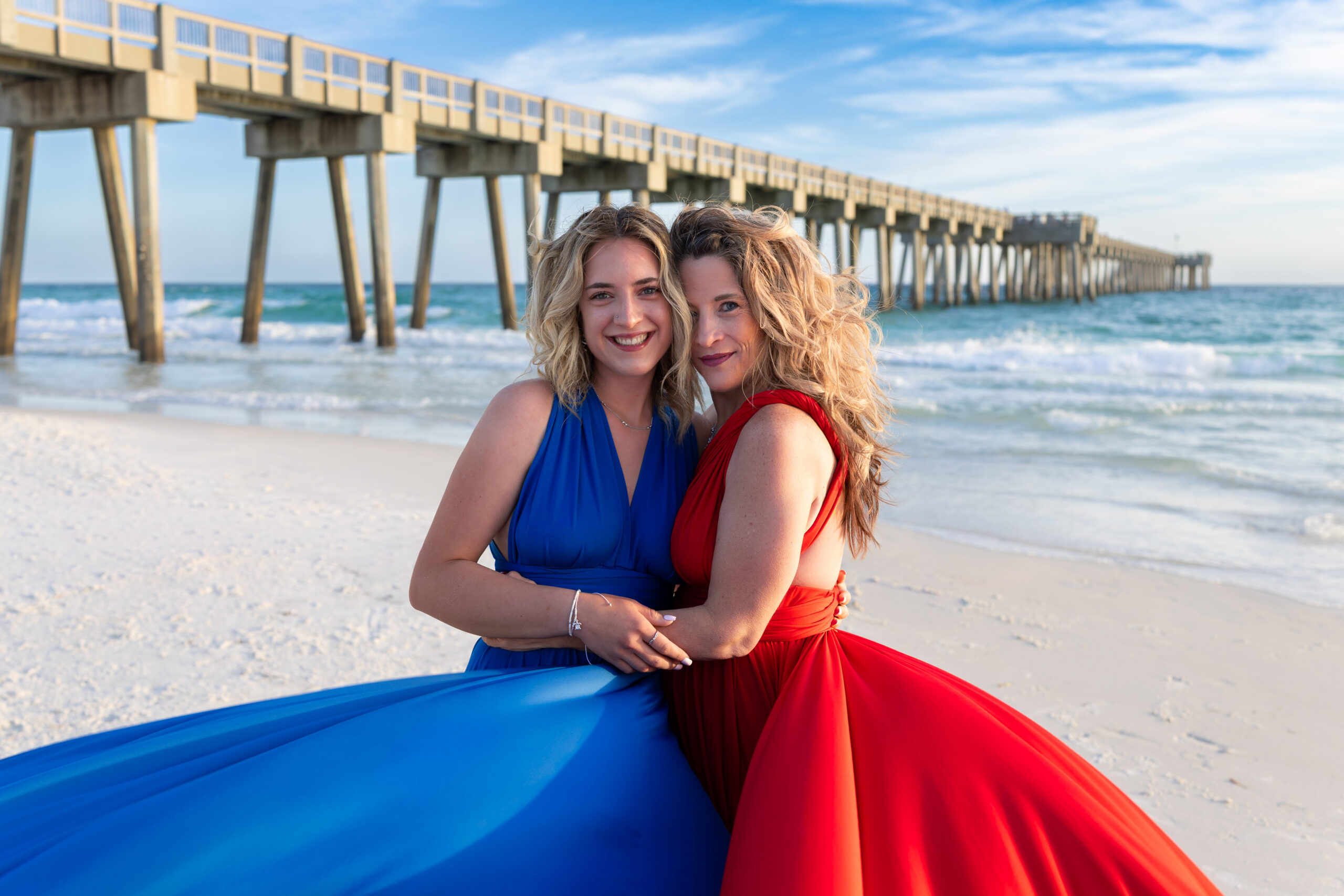 IMG_6407 Two friends embracing by a beach pier in blue and red flying dresses
