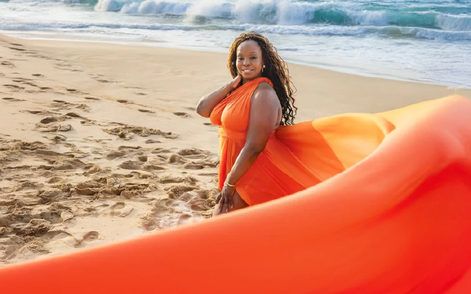 Woman in an orange flying dress sitting on a sandy beach with waves behind her