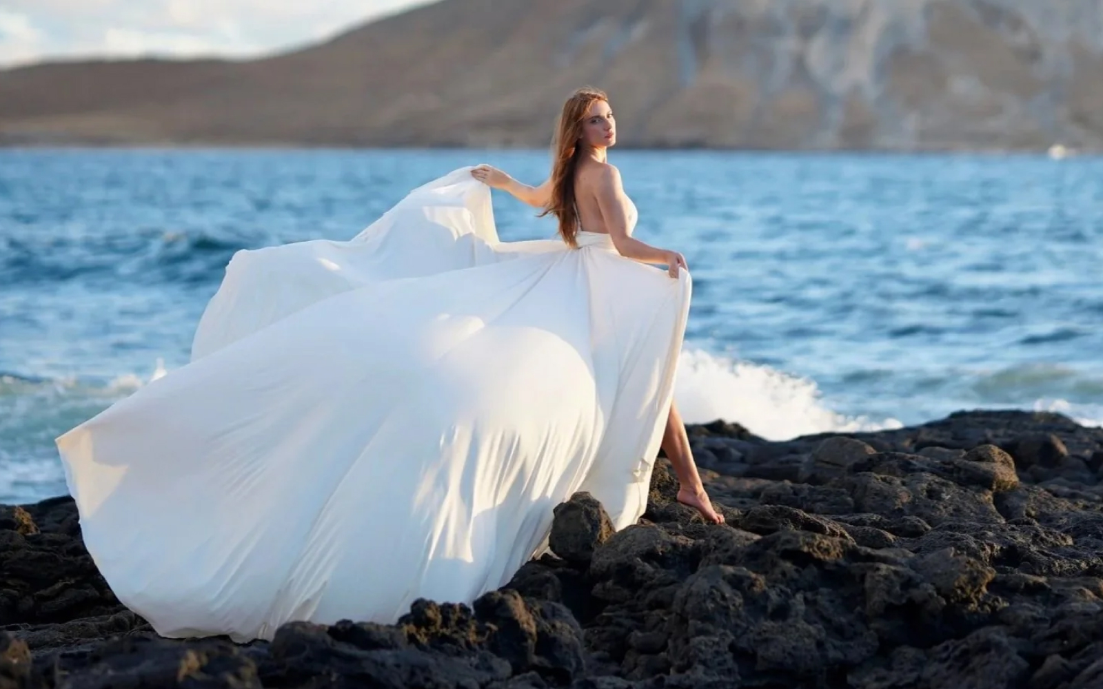 Woman in a white flying dress on dark lava rocks by the sea